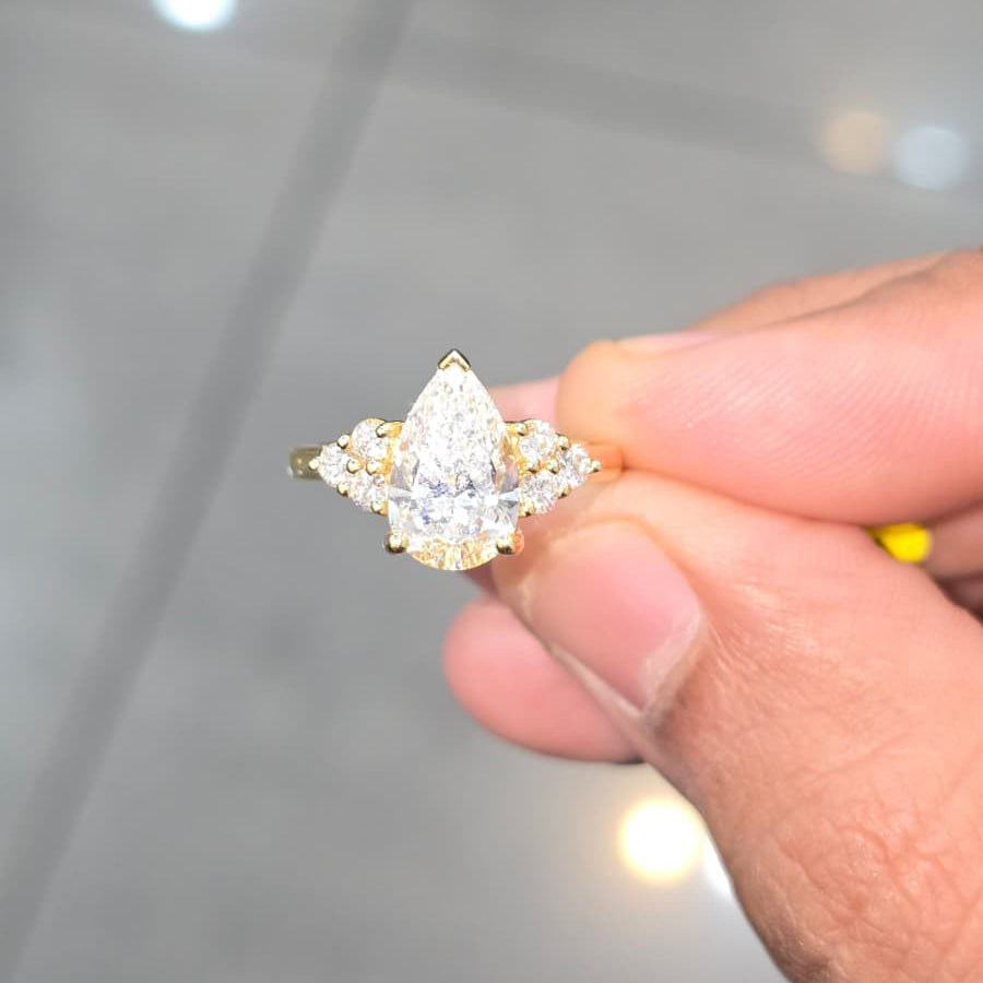 A close-up of a hand holding a small pear-cut diamond ring with gold and silver accents against a blurred gray tiled background.